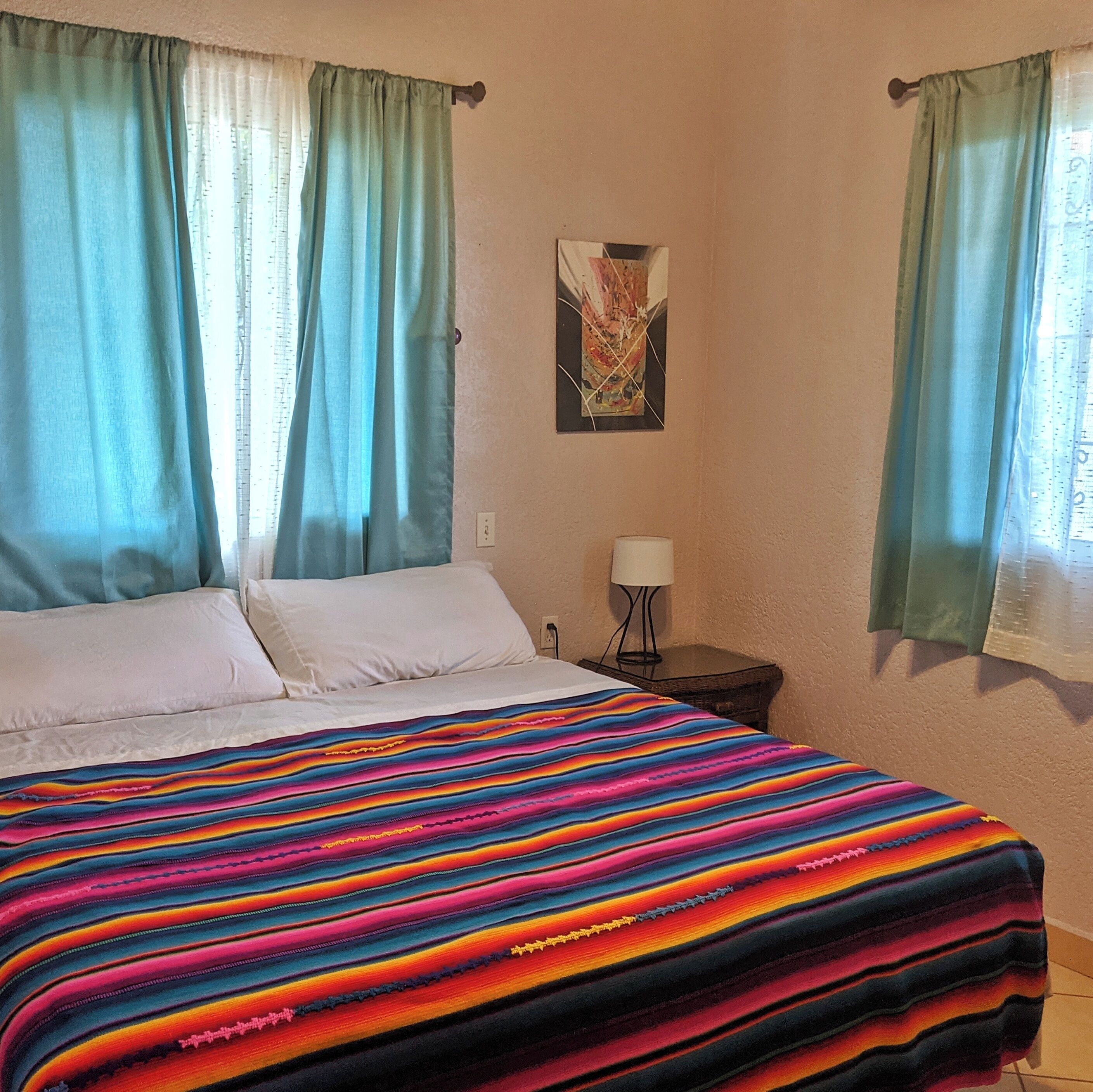 A bedroom with a bed, two pillows, a colorful striped blanket, a nightstand with a lamp, and a painting on the wall.