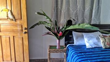 Una habitación con puerta de madera, cama con colcha azul a rayas, una pequeña mesita de mimbre con una planta y una cortina estampada con motivos paisley.