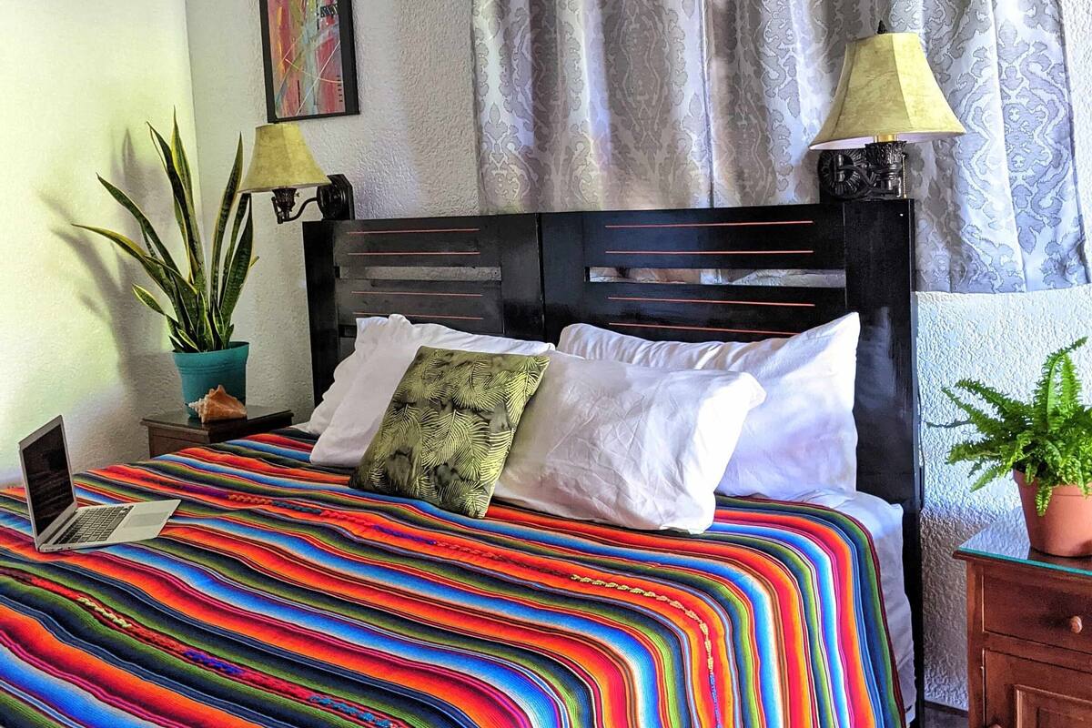 A bedroom with a bed, two lamps, a potted plant, and a painting on the wall.