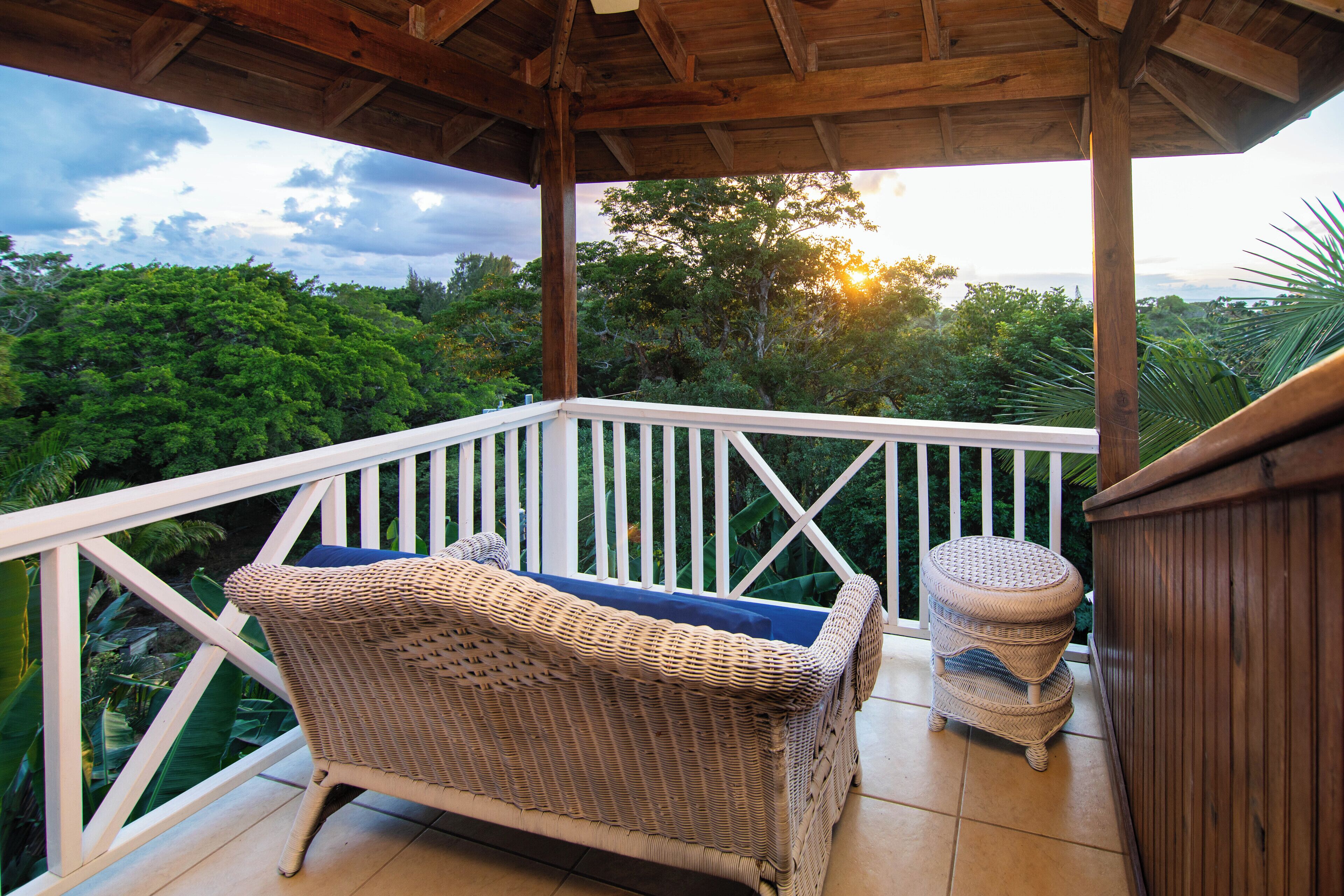 A wicker chair with a cushion on a balcony with a white railing, overlooking a lush green landscape.