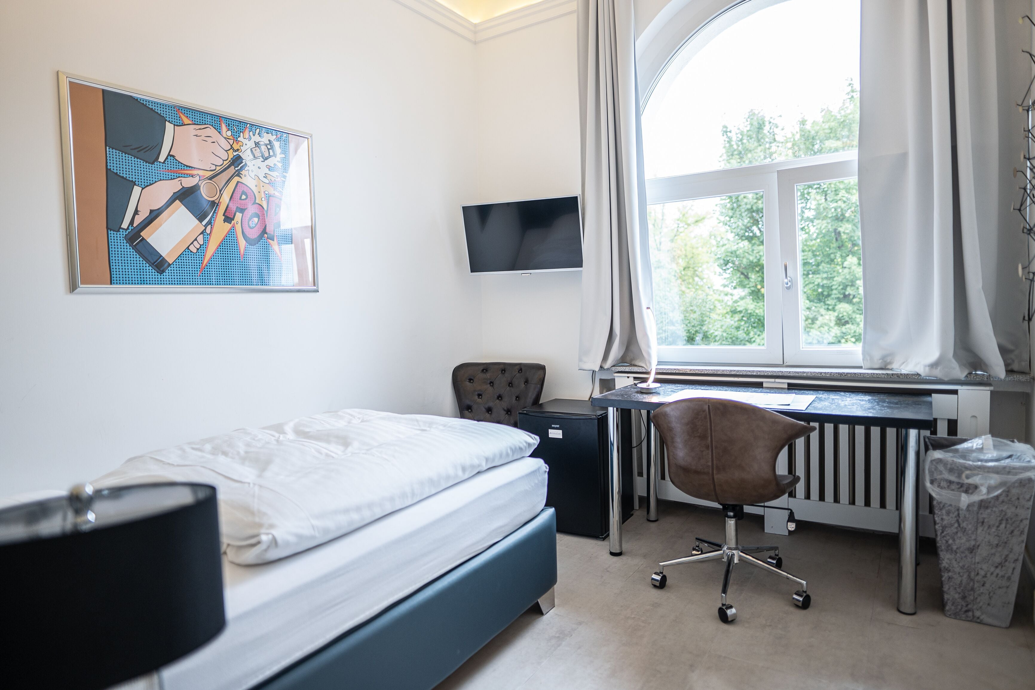Quarto com cama, escrivaninha, cadeira, televisão, radiador e janela com cortinas.