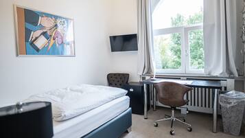Quarto com cama, escrivaninha, cadeira, televisão, radiador e janela com cortinas.