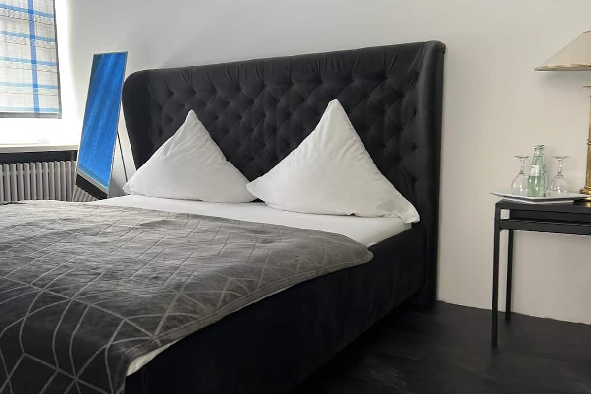 A neatly made bed with a black headboard and white pillows, a bedside table with a lamp, and a window with blue curtains.