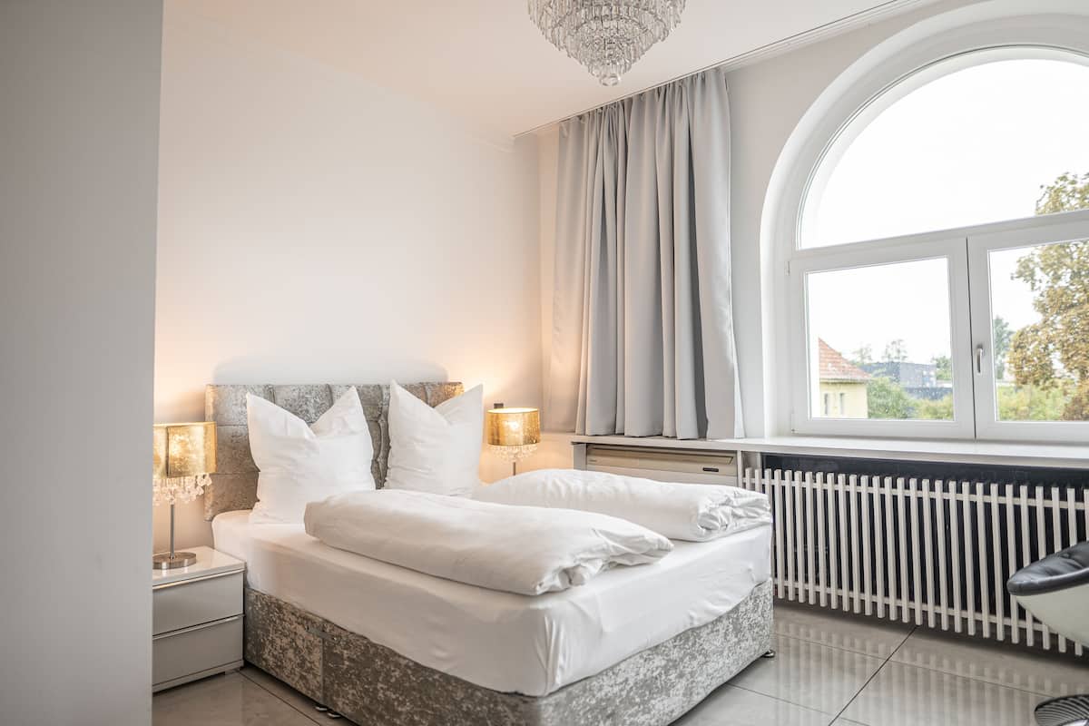 A bedroom with a large window, a bed with white linens, a nightstand with a lamp, and a radiator.