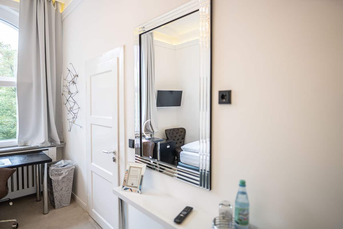A hotel room with a large mirror, a bed, a desk, a chair, a TV, a trash bin, and a water bottle.