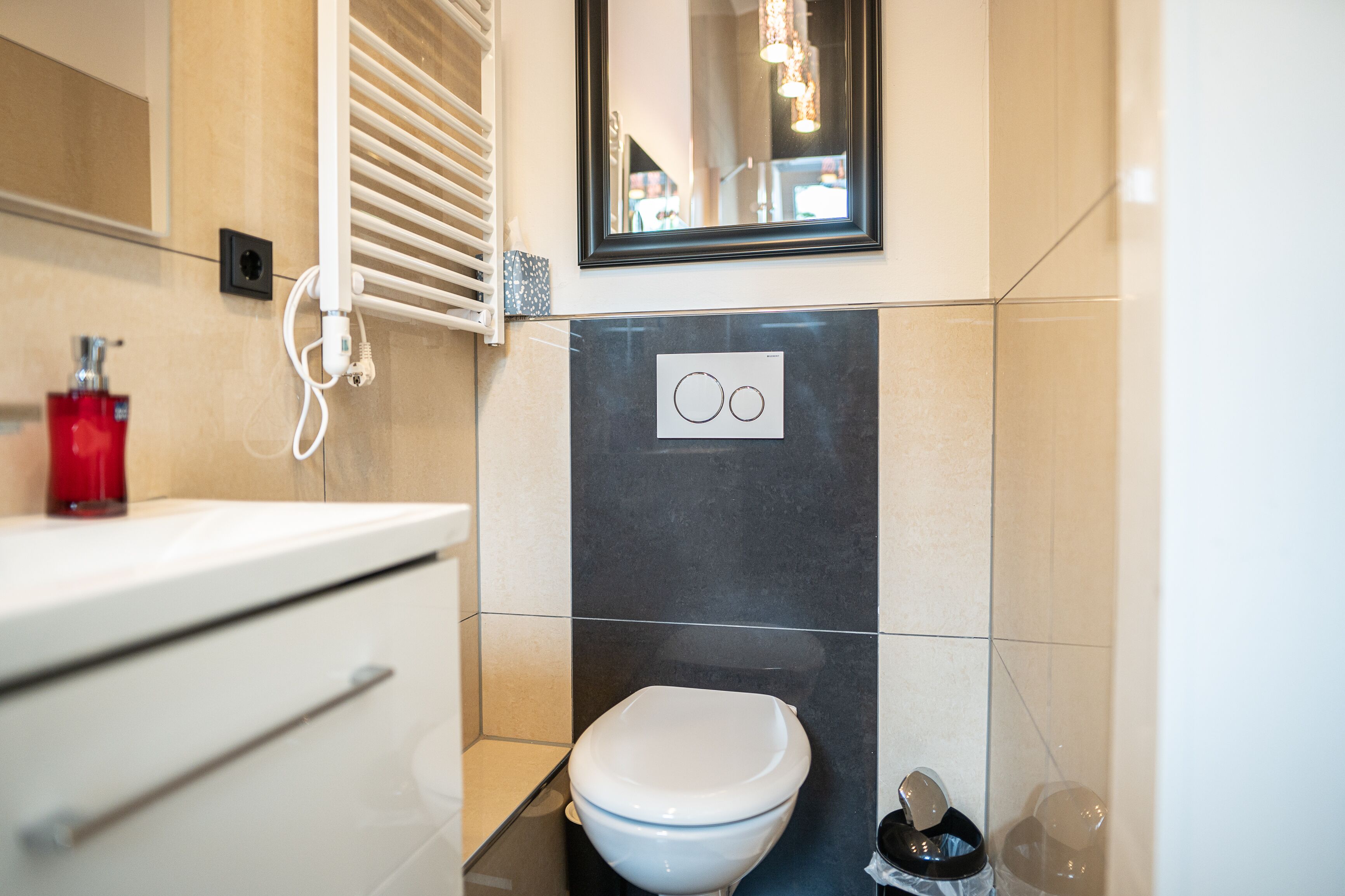 Un baño moderno con inodoro blanco, dispensador de jabón rojo y espejo sobre el lavabo.