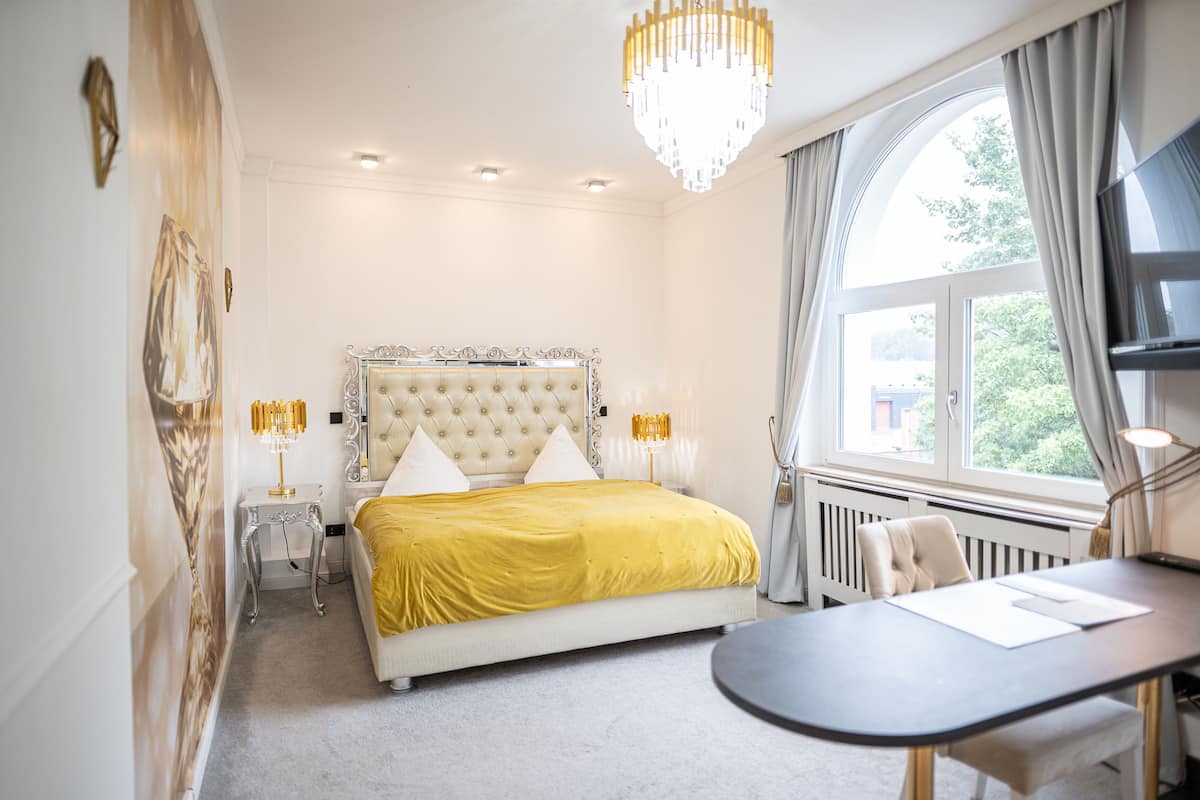 A bedroom with a large bed, a bedside table, a chair, a desk, and a large window.