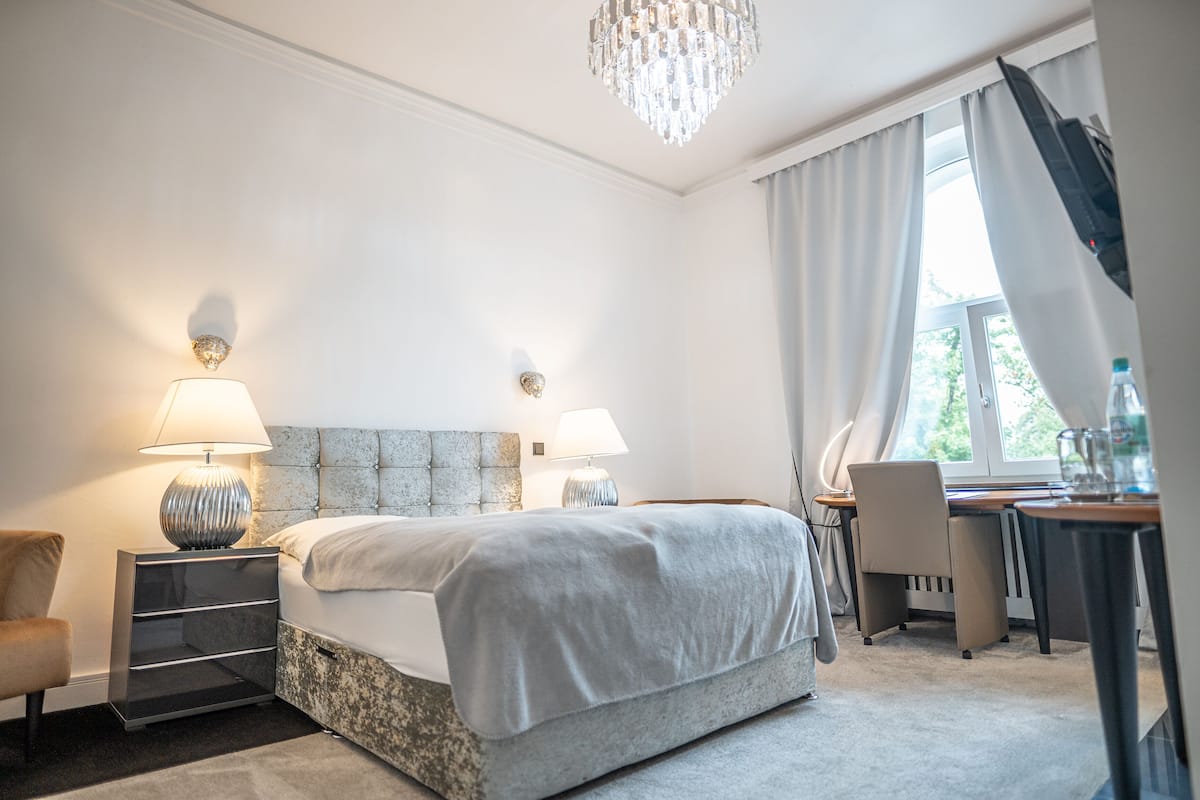 A hotel room with a large bed, two bedside lamps, a chandelier, a desk with a chair, and a window with curtains.