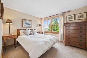 Quarto com cama, mesas de cabeceira, cômoda, janela com cortinas e quadros nas paredes.