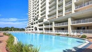 A large swimming pool surrounded by a landscaped area with plants, a multi-story building with balconies, and a clear blue sky. - ☀️By Beach in Seascape-18th FL Gulf Views-3 Pools+Sauna-2BR Ariel Dunes II 1807 (Miramar Beach)