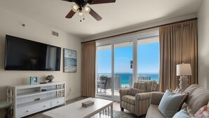 A spacious living room with a large flat-screen TV, a ceiling fan, a sofa, a coffee table, and a view of the ocean through large sliding doors. - ☀️By Beach in Seascape-18th FL Gulf Views-3 Pools+Sauna-2BR Ariel Dunes II 1807 (Miramar Beach)