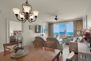 A spacious living area with a dining table, chairs, a chandelier, a ceiling fan, and a large window offering a view of the ocean. - ☀️By Beach in Seascape-18th FL Gulf Views-3 Pools+Sauna-2BR Ariel Dunes II 1807 (Miramar Beach)