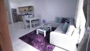 Una sala de estar moderna con un sofá blanco, una mesa de centro de vidrio y un tapete morado. Incluye una zona de cocina con gabinetes blancos y una mesa de comedor con sillas.