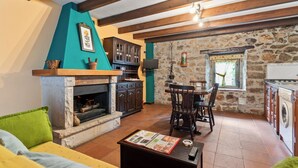 Un salon chaleureux avec une cheminée, des murs en pierre, des poutres en bois, un coin repas et une cuisine équipée d’un lave-linge.