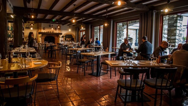 Un restaurant amb diverses taules i cadires, una llar de foc de maó i grans finestres.