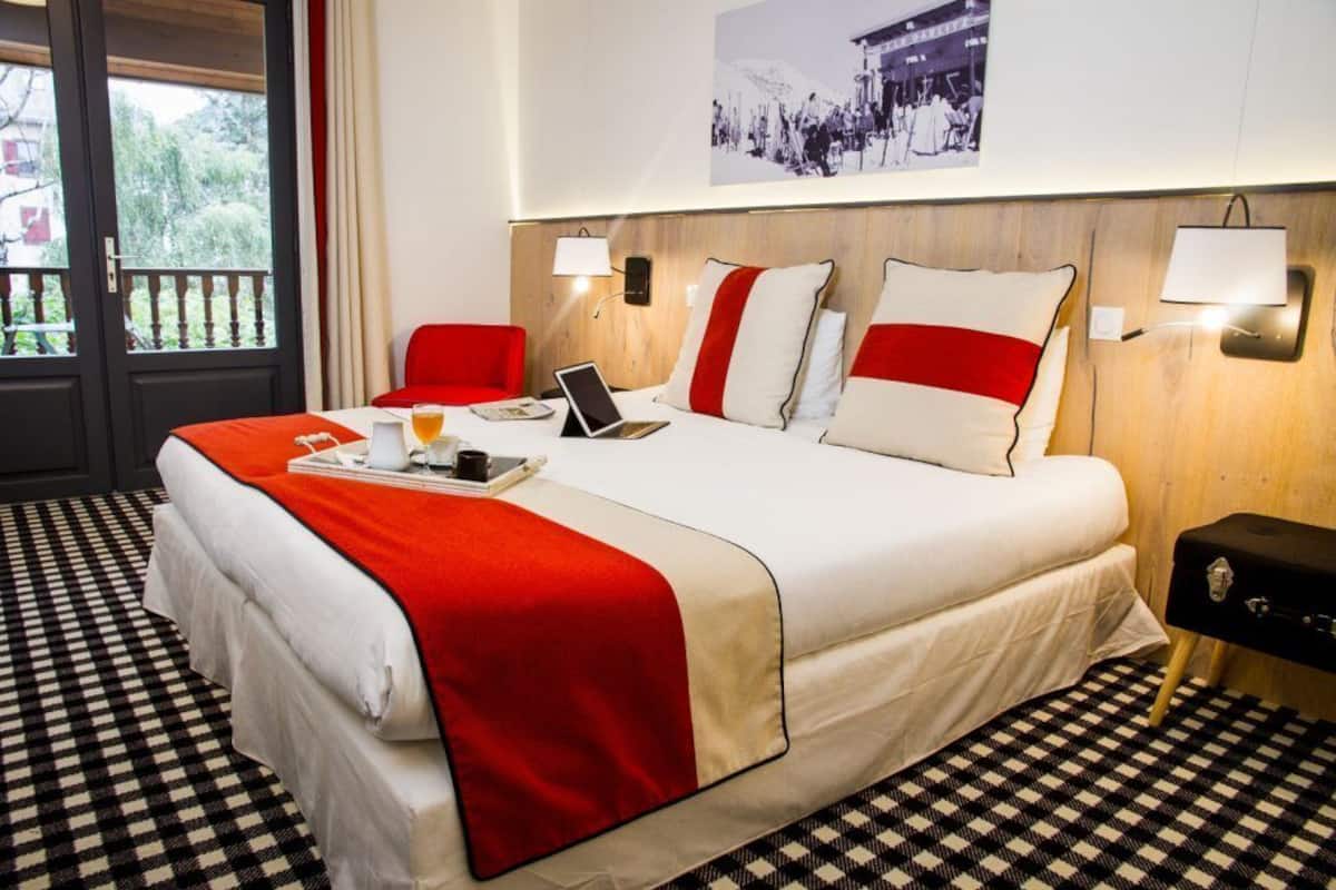 Ein Hotelzimmer mit Schachbrettmuster-Boden, einem Bett mit rotem und weißem Überwurf, zwei roten Stühlen, einem Laptop auf dem Bett und einem kleinen Tisch mit einer Tablett voller Gegenstände.