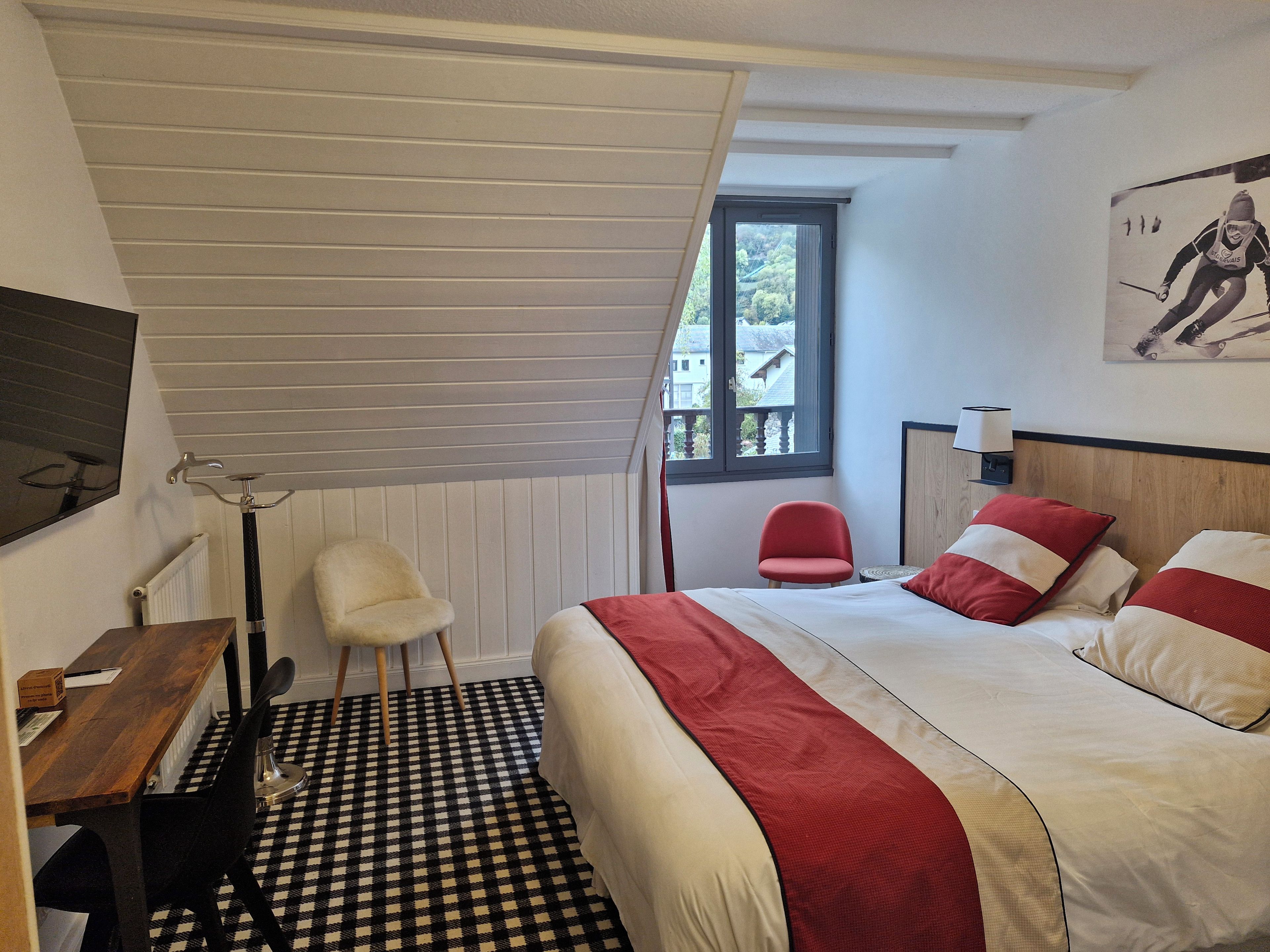 A bedroom with a checkered floor, a bed with red and white bedding, a desk with a chair, a window with a view, and a wall-mounted lamp.