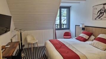 A bedroom with a checkered floor, a bed with red and white bedding, a desk with a chair, a window with a view, and a wall-mounted lamp.