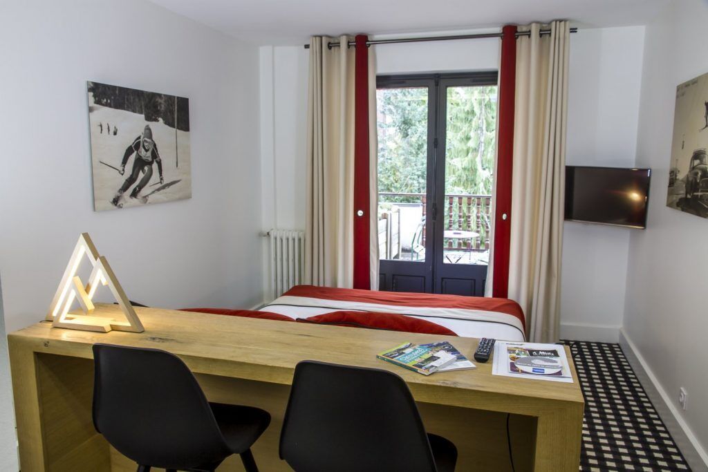 A hotel room with a bed, a desk with two chairs, a TV, and a window with curtains.