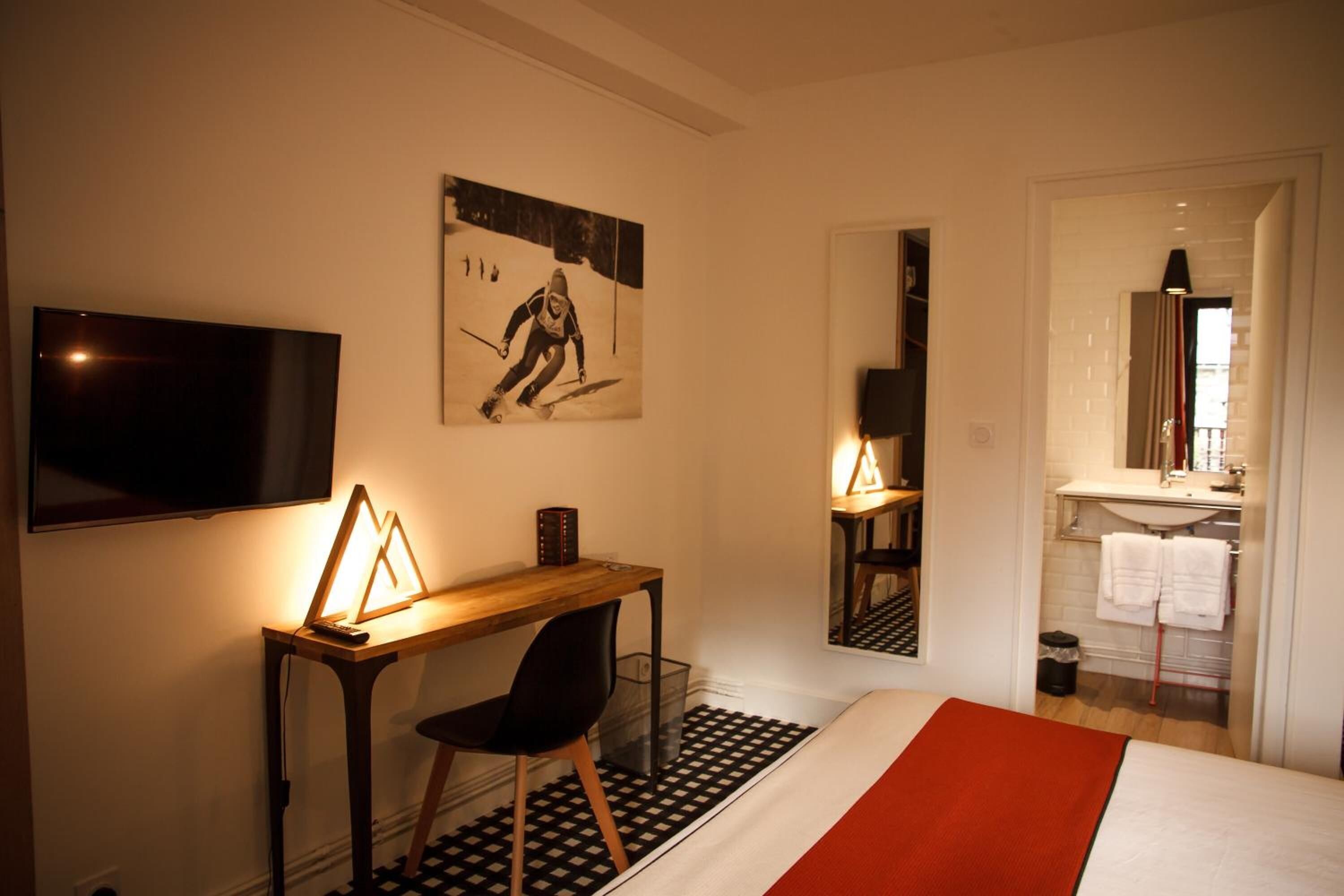 A hotel room with a bed, a desk with a chair, a television, a painting on the wall, and a bathroom visible through an open door.