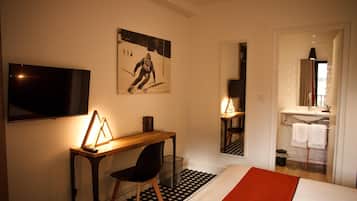 A hotel room with a bed, a desk with a chair, a television, a painting on the wall, and a bathroom visible through an open door.
