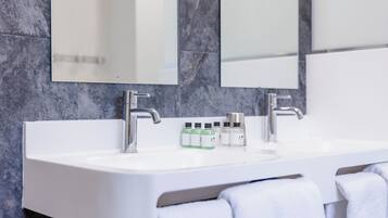 Ein modernes Badezimmer mit einem weißen Waschbecken, zwei Wasserhähnen und einem Spiegel darüber. Auf dem Waschbecken stehen Flaschen mit Toilettenartikeln.