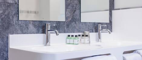 A modern bathroom with a white sink, two faucets, and a mirror above. There are bottles of toiletries on the sink.
