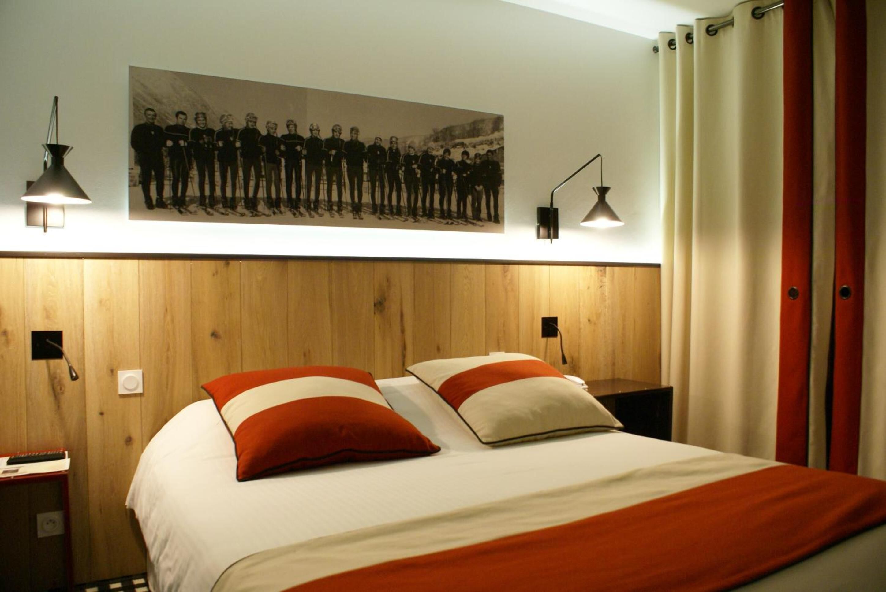 A hotel room with a wooden headboard, two bedside tables, a bed with white and orange bedding, and a large group photo on the wall.