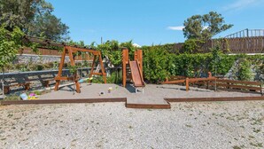 A playground with wooden swings and a slide, surrounded by a gravel area and a wooden fence. - Holiday home in Kiotari with a terrace (Kiotari)