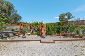 A playground with wooden swings and a slide, surrounded by a gravel area and a wooden fence.