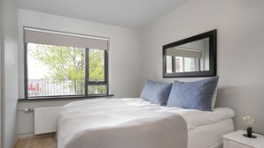 A bedroom with a large window, a bed with white bedding, a bedside table with a lamp, and a mirror on the wall. - Downtown Apartment by Old Harbor (Reykjavík)