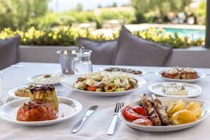 Ein Tisch ist reichhaltig mit Speisen gedeckt, darunter gefülltes Gemüse, Salat und gegrilltes Fleisch. Im Hintergrund sieht man einen Pool und Grünflächen.