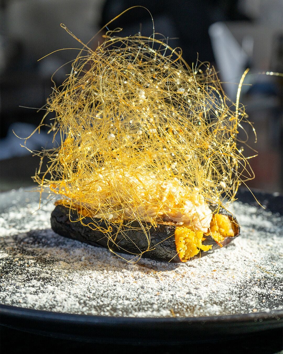 A dish with a golden, crispy texture, possibly a fried item, on a dark plate with a textured surface.