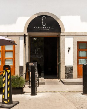 Entrada de un hotel con un letrero que dice 'CASA DE LA LUZ HOTEL BOUTIQUE', el número '7' en la pared y una barrera de malla metálica.