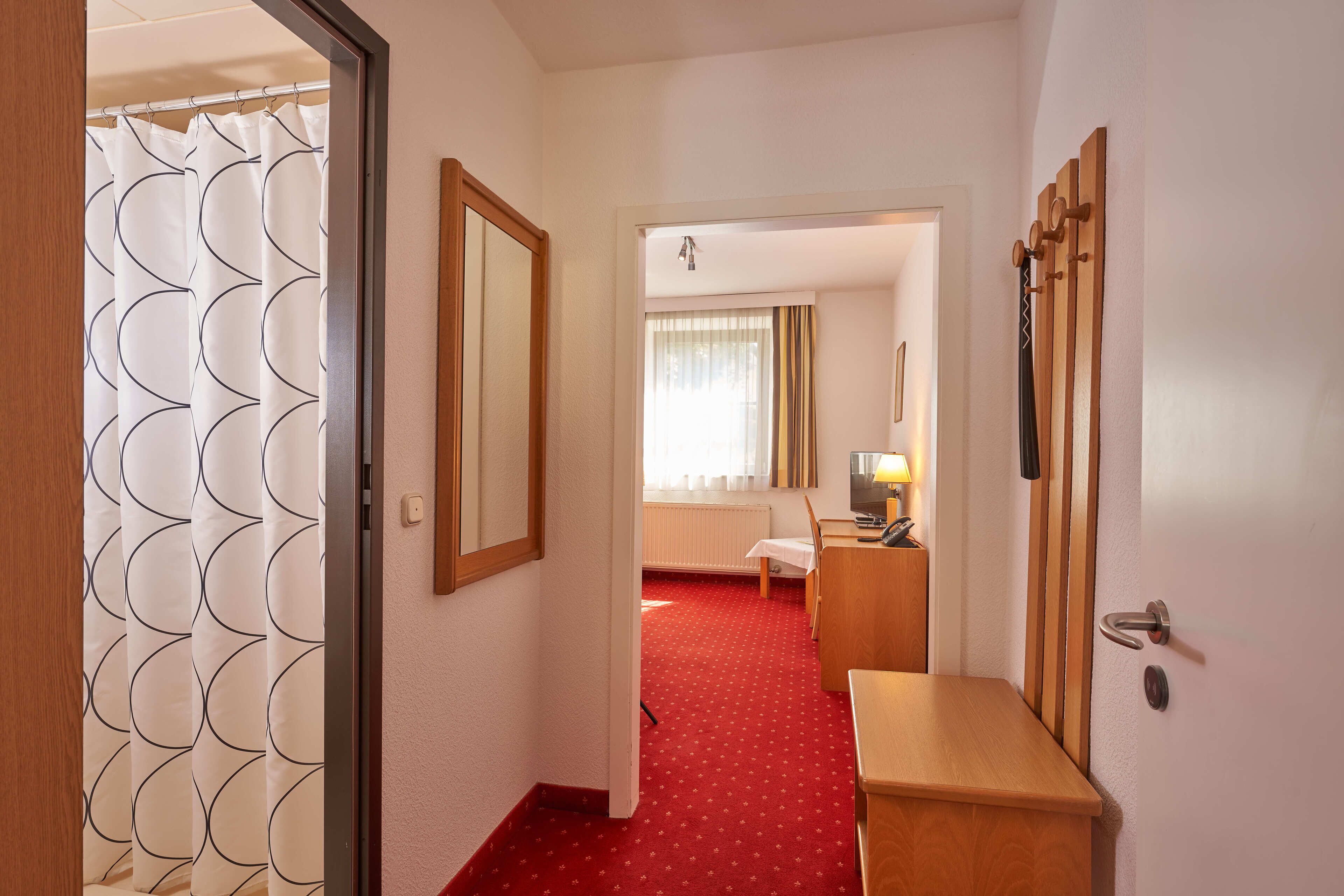 Ein Hotelgang mit rotem Teppich, einem Holztisch, einem Spiegel und einer Tür zu einem Zimmer mit Bett und Fenster.