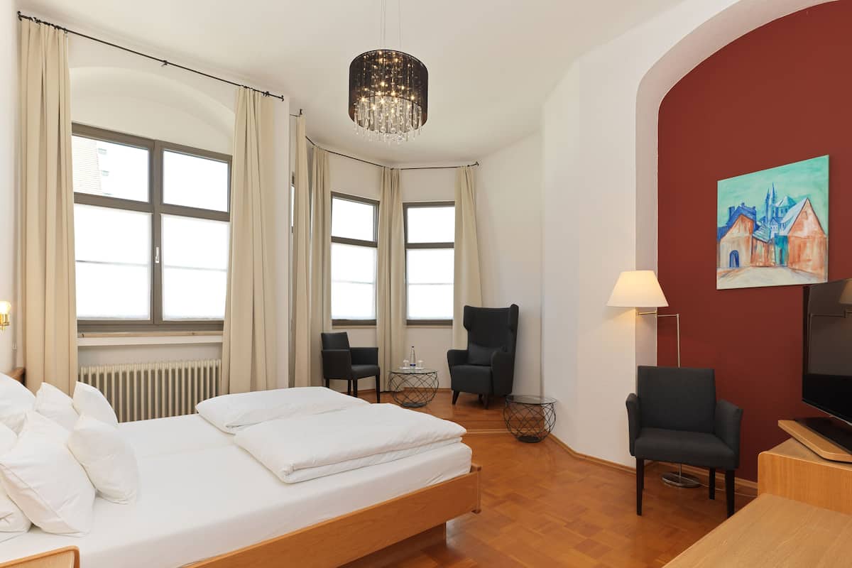 A hotel room with a bed, two armchairs, a small table, a TV, and a painting on the wall.