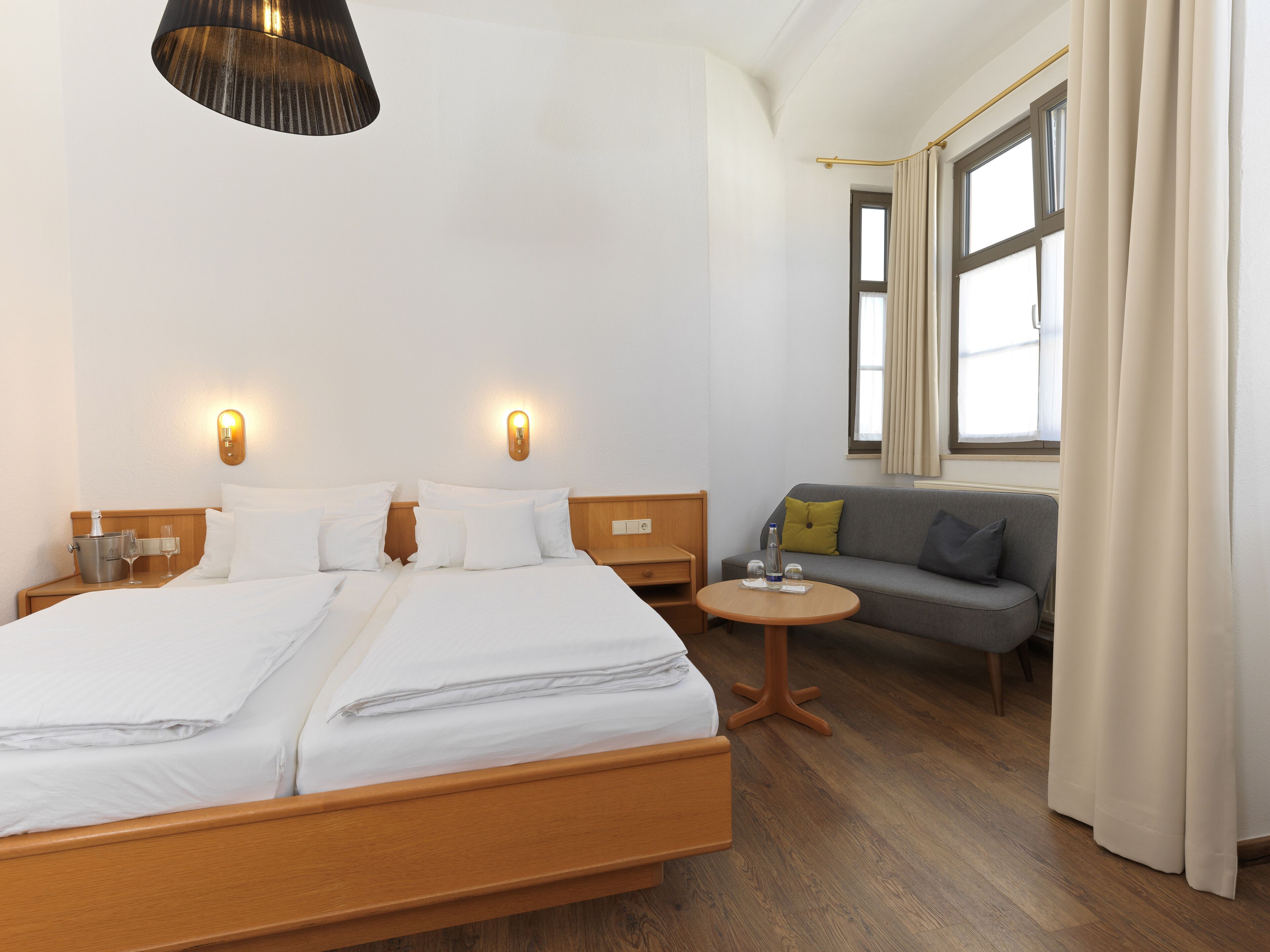 Habitación de hotel con una cama grande, mesitas de noche, un sofá, una pequeña mesa redonda y una ventana con cortinas.