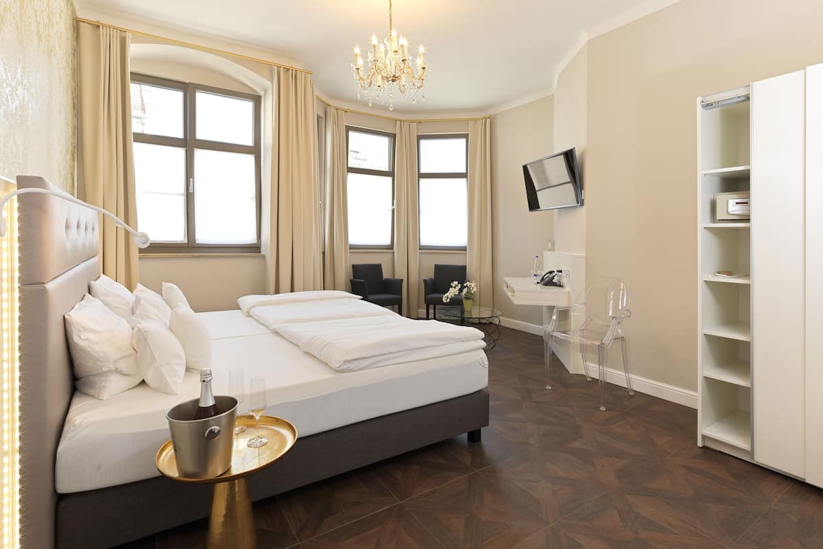 A modern hotel room with a large bed, a desk, a chair, a TV, and a minibar.