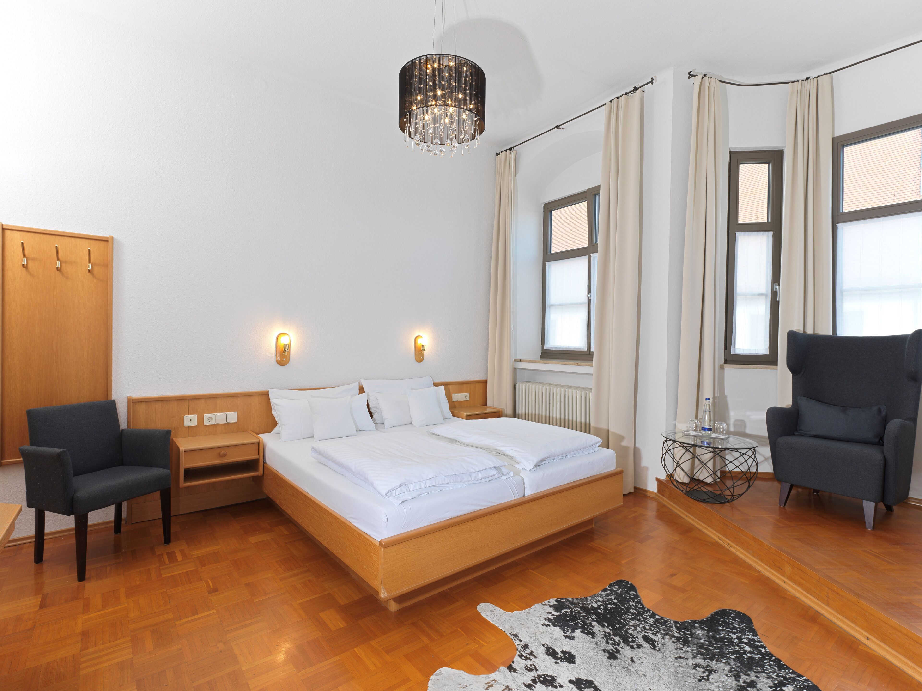 Una habitación de hotel moderna con una cama de madera, dos mesitas de noche, una araña, un ventanal con cortinas y una mesa de centro redonda, además de dos sillones.