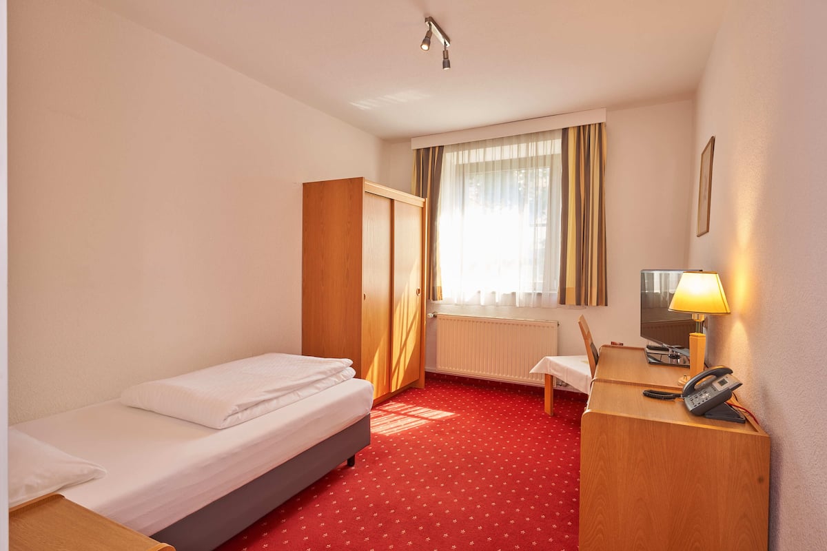 A hotel room with a single bed, a desk with a telephone, a chair, a television, and a window with curtains.