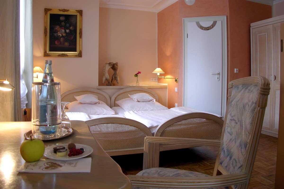 A hotel room with two beds, a dining table with fruit and a bottle, a painting on the wall, and a door with a decorative wreath.