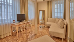 A hotel room with a wooden herringbone floor, a beige sofa, a TV on a wooden desk, and sheer curtains. - Lodner Genießerhotel Drei Mohren (Lauingen)