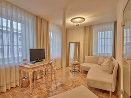 A hotel room with a wooden herringbone floor, a beige sofa, a TV on a wooden desk, and sheer curtains.