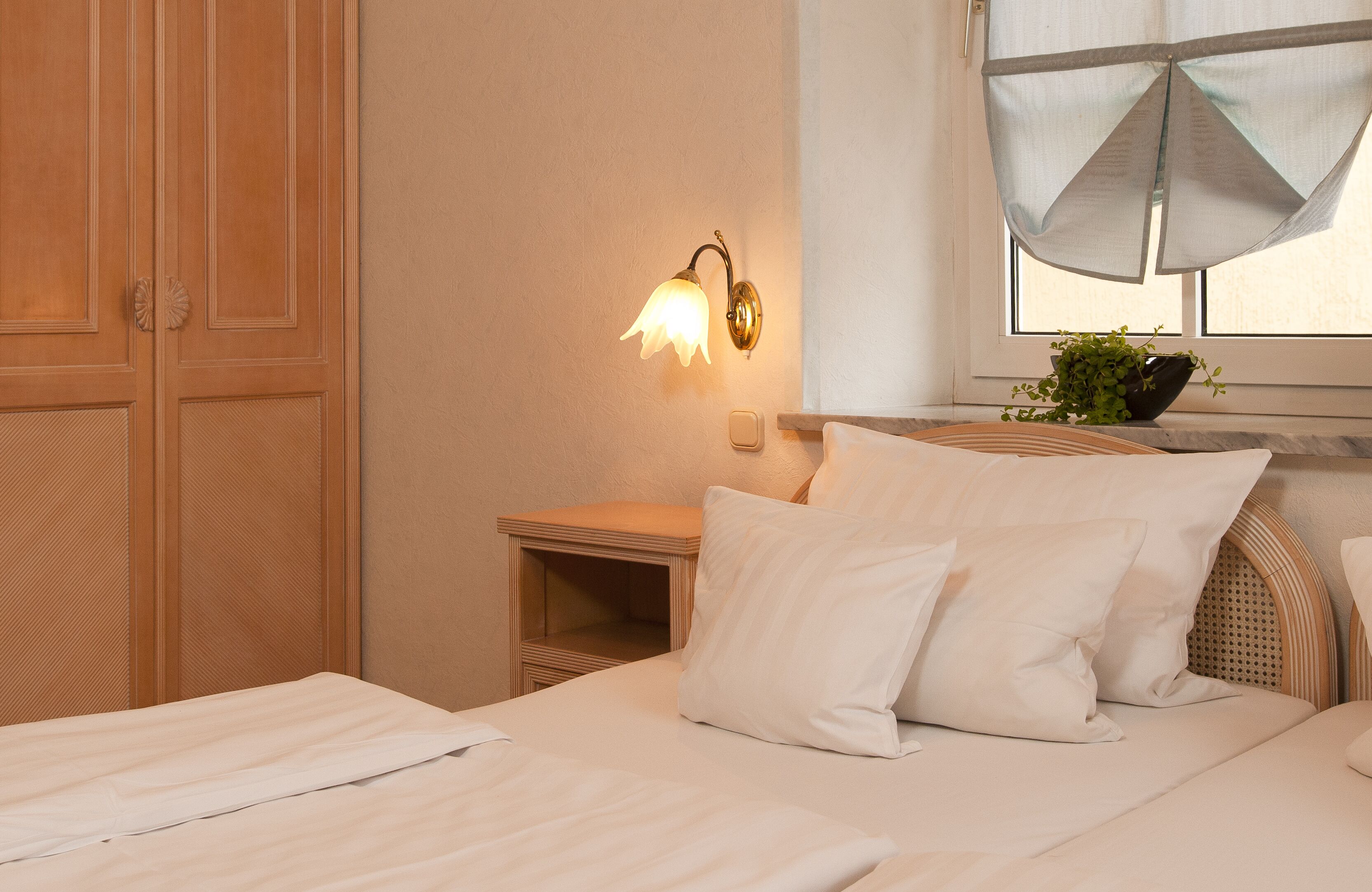 Una habitación de hotel con una cama, almohadas, una mesita de noche, una lámpara de pared, una ventana con cortina translúcida y una planta en maceta.