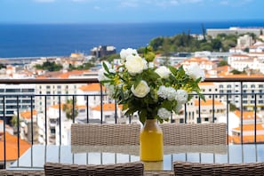 Un balcón con mobiliario de mimbre, una mesa de vidrio y un jarrón amarillo con flores blancas.