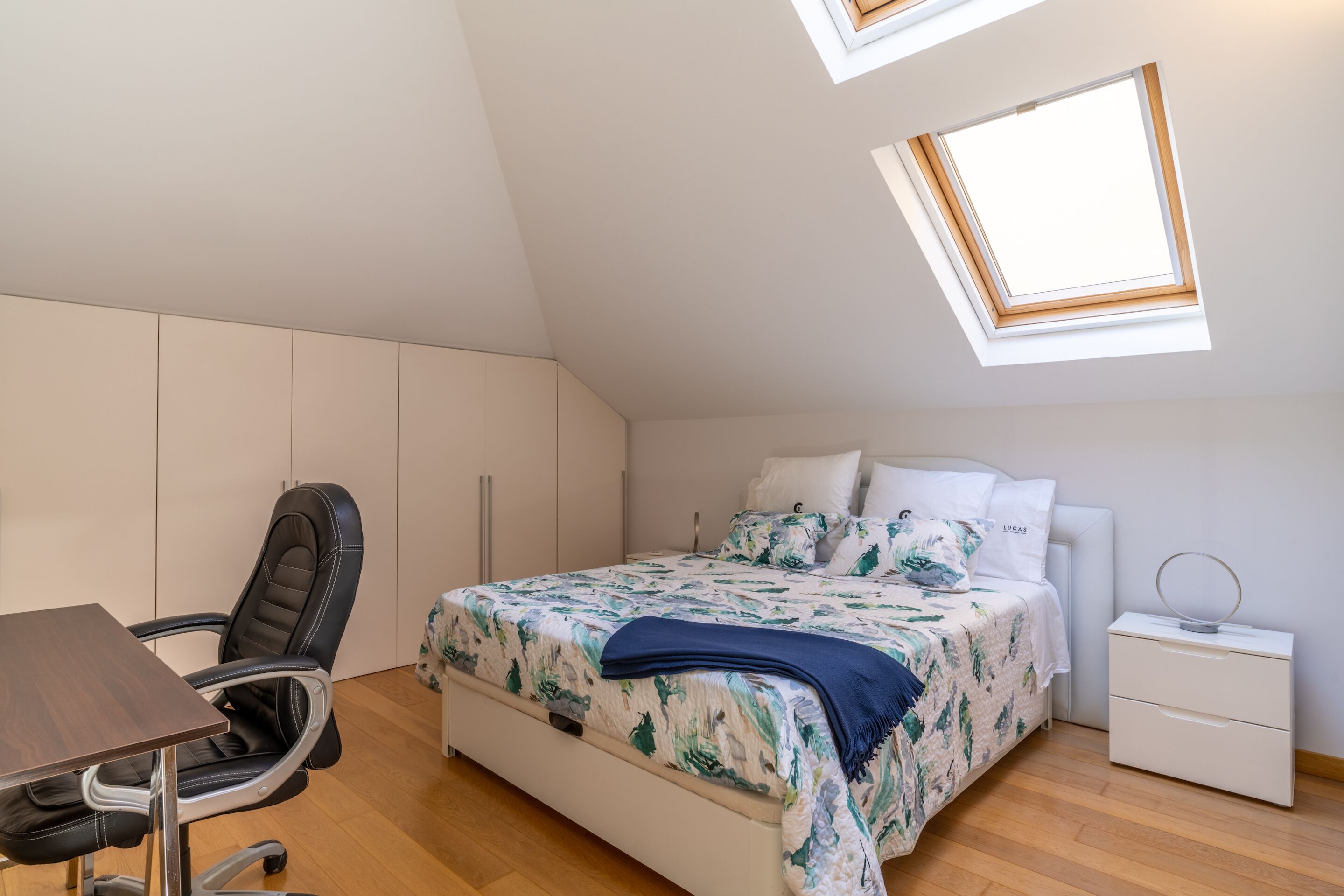 A bedroom with a bed, a desk, a chair, a nightstand, and a skylight.