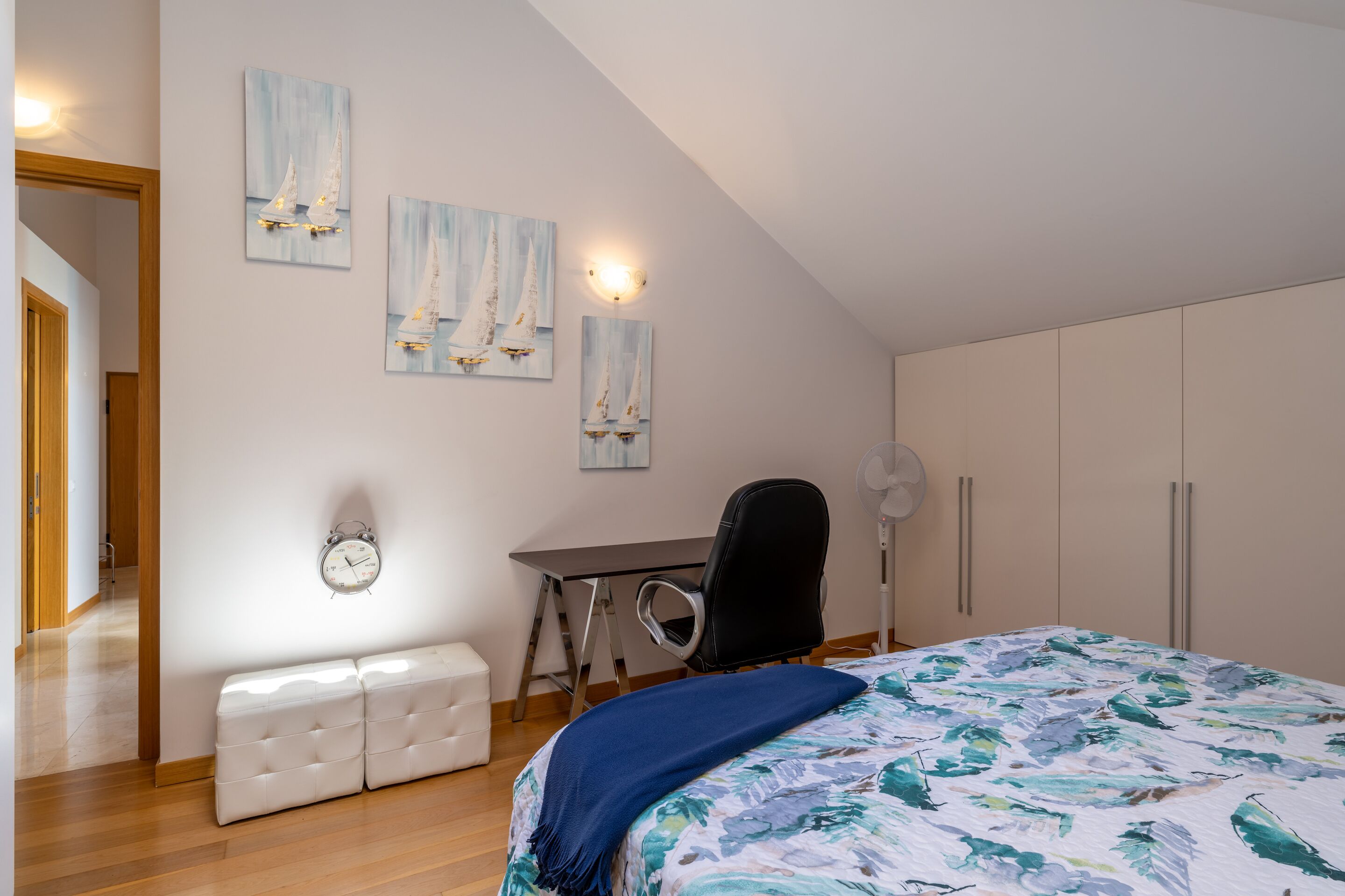 A bedroom with a bed, a desk, a chair, a bench, and wall-mounted artwork.