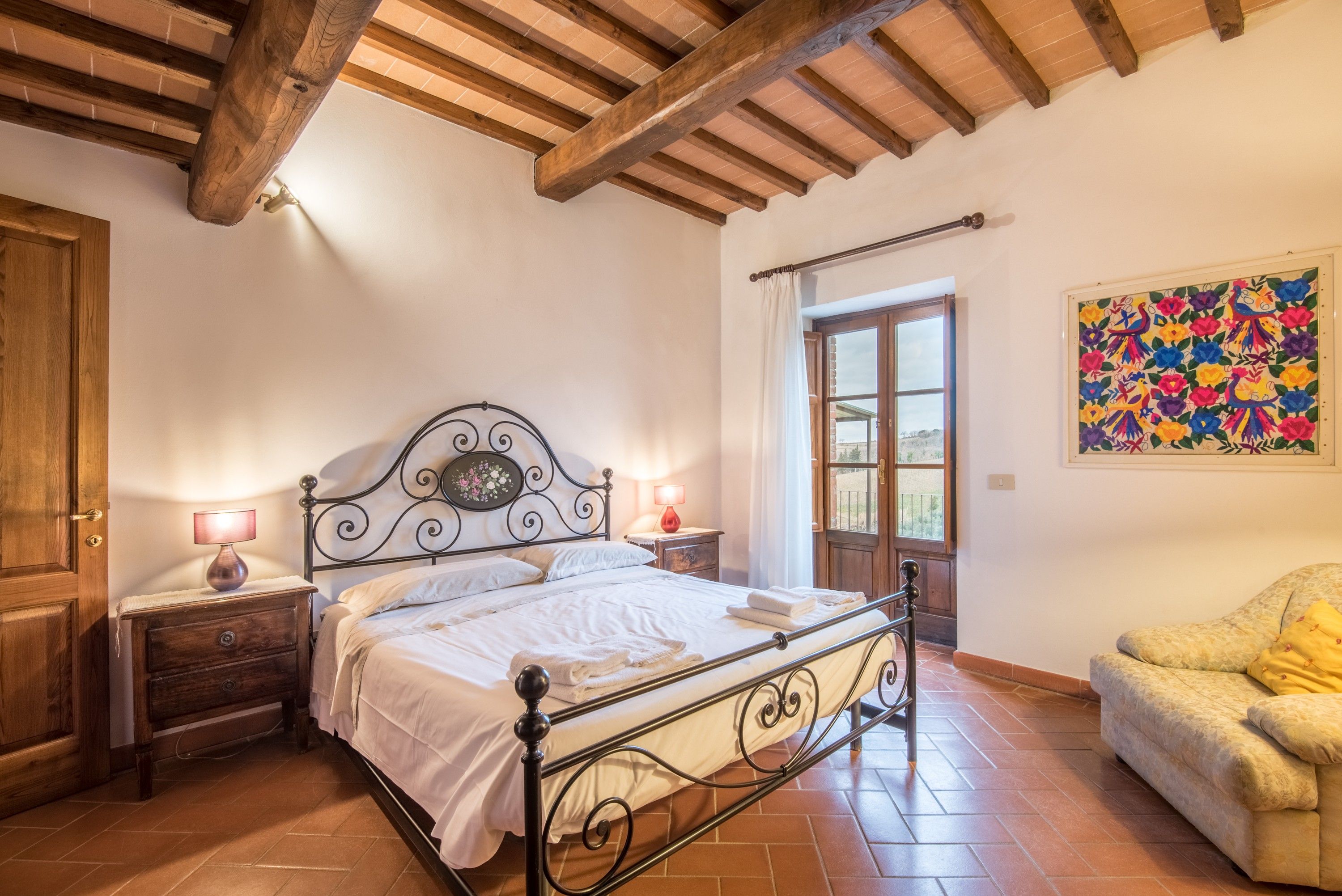 A bedroom with a bed, nightstands, a wooden door, a window, and a colorful wall decoration.