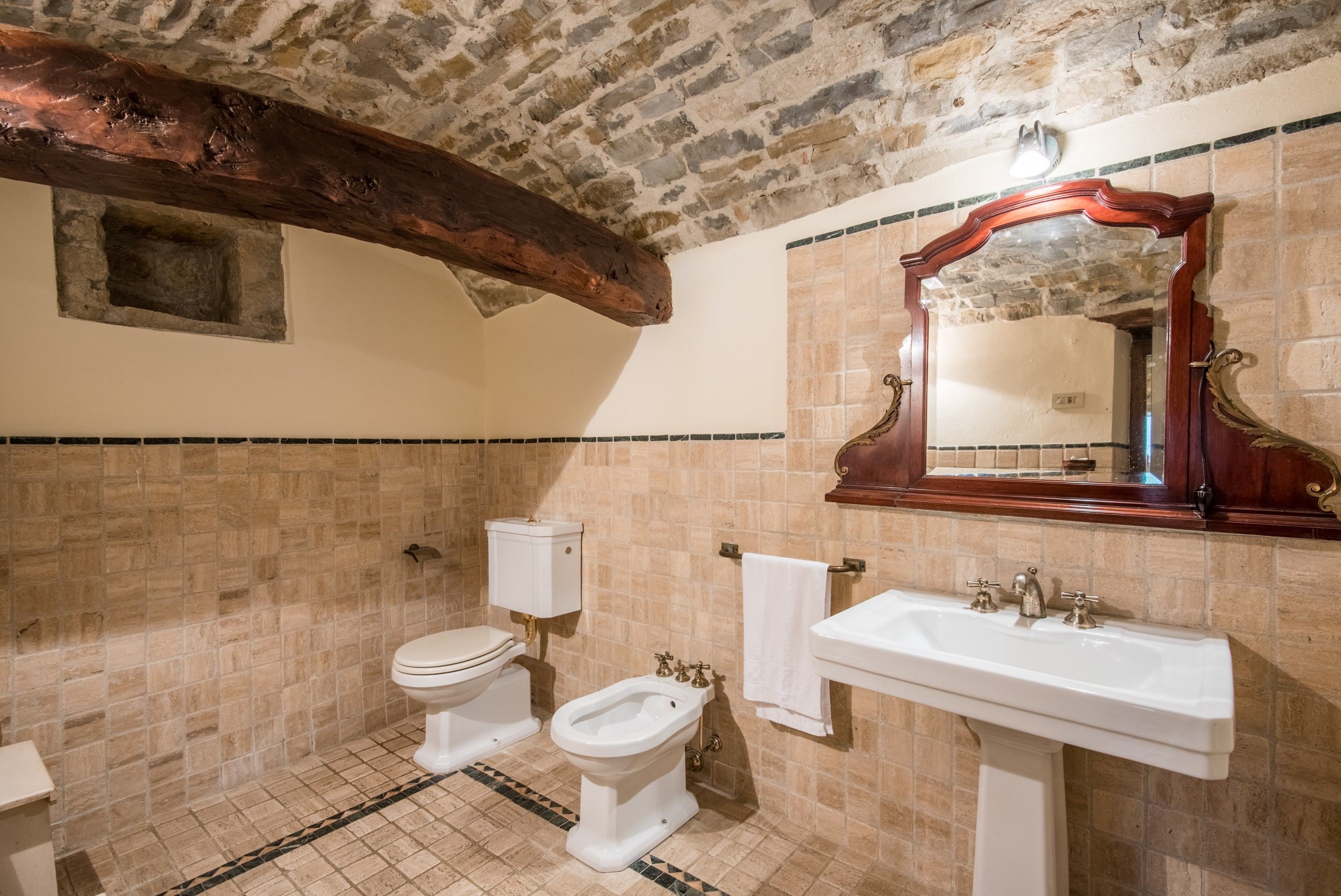 A bathroom with a stone wall, a wooden ceiling beam, a white toilet, a bidet, and a sink with a mirror above it.