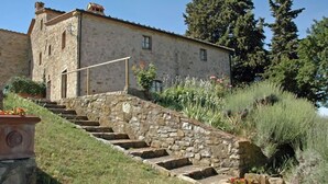 Una scala in pietra conduce a un edificio rustico in pietra con tetto piatto, immerso nel verde e alberi.